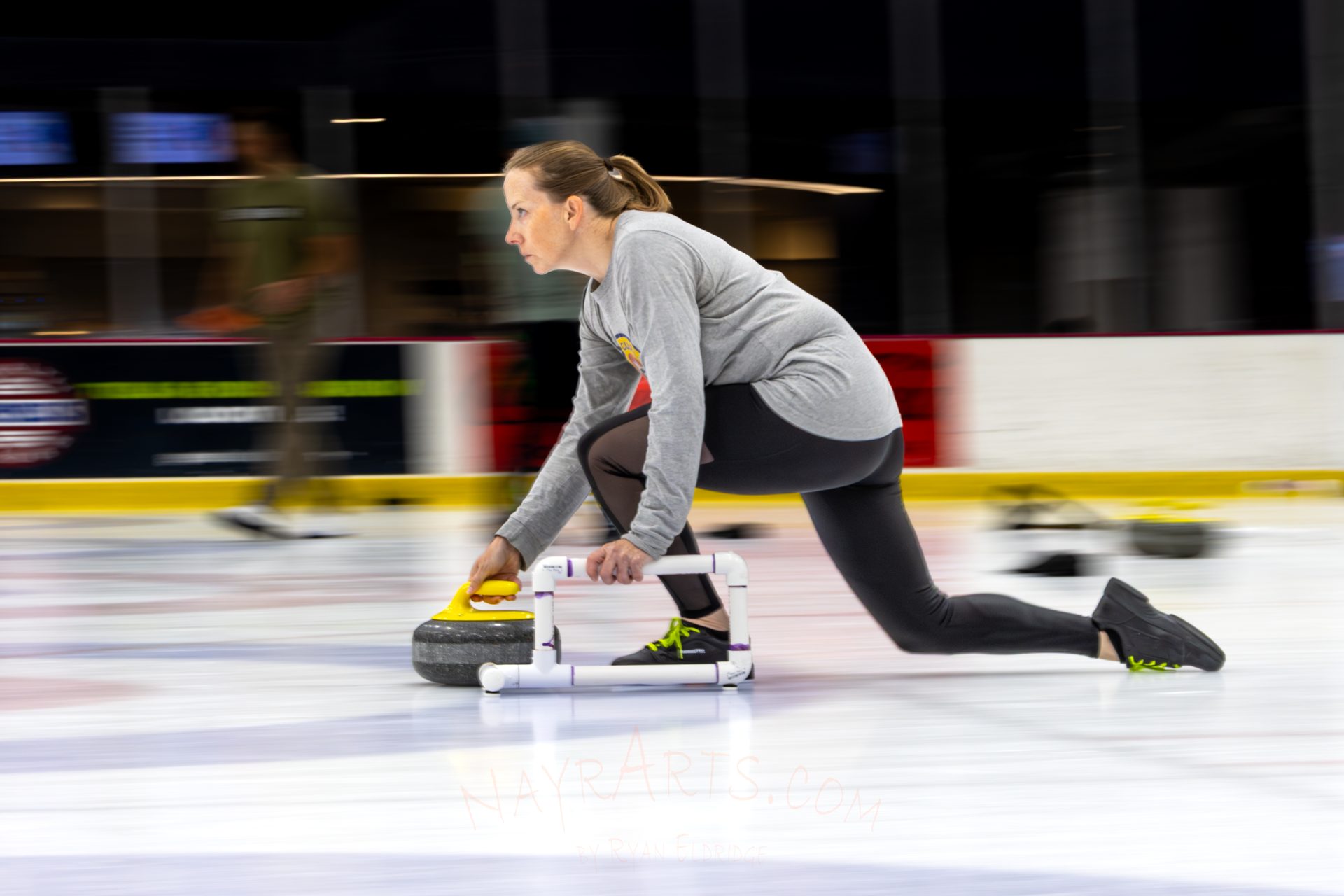 A curler in a grey club shirt slides low on the ice, guiding a yellow-handled stone forward
